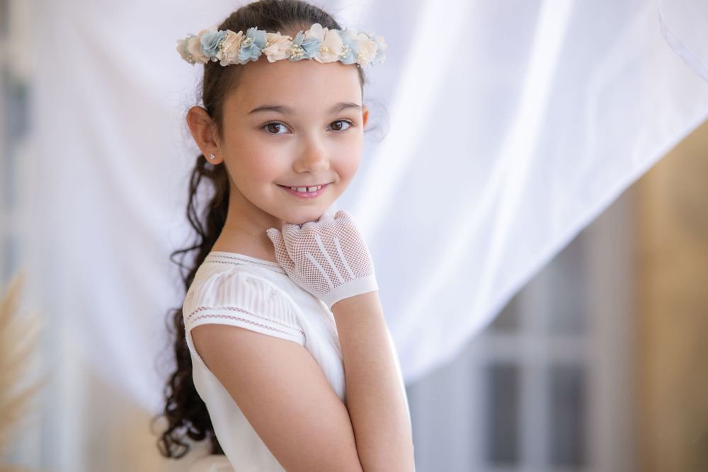 Young girl in white dress poses gracefully against white curtain backdrop during indoor photo session.