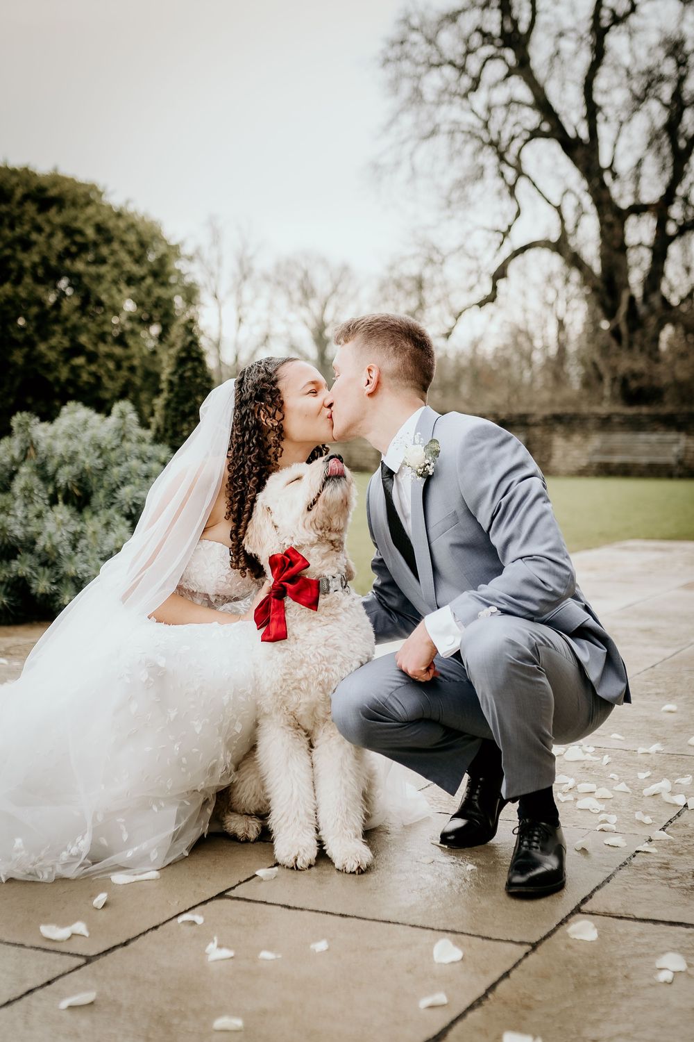 Wedding couple shares intimate moments with their fluffy white dog on stone patio with scattered petals.