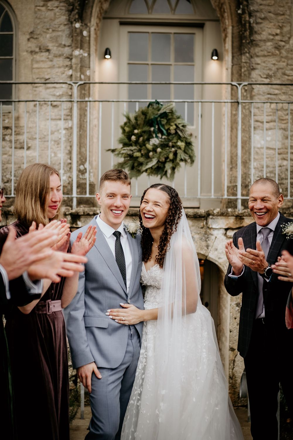 Wedding party celebrates with joyful laughter and applause against a rustic stone wall backdrop.