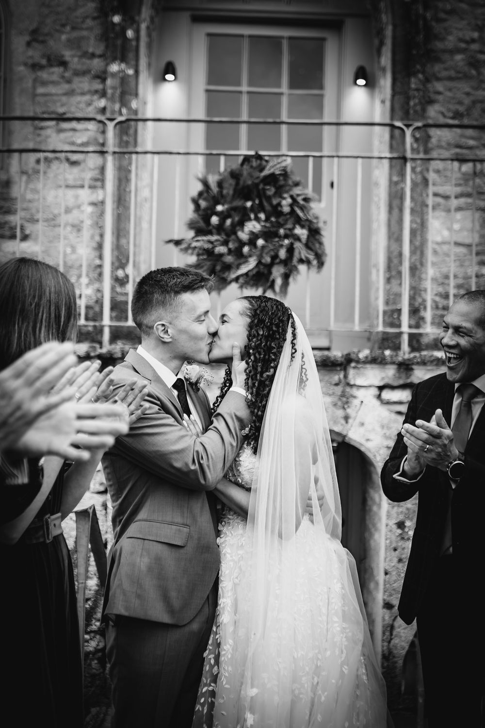 Newlyweds share their first kiss outside a stone building with a decorative wreath while guests celebrate.