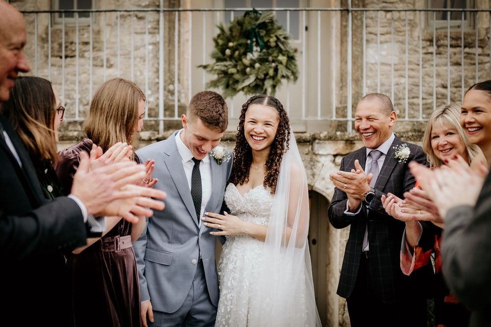 Wedding celebration scene with guests clapping and cheering outside a stone building.