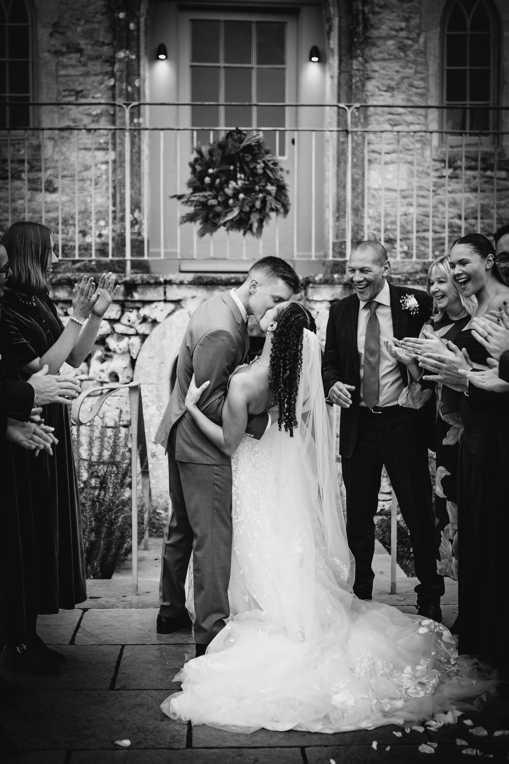 A romantic wedding kiss captured in black and white with guests applauding at a historic stone venue.