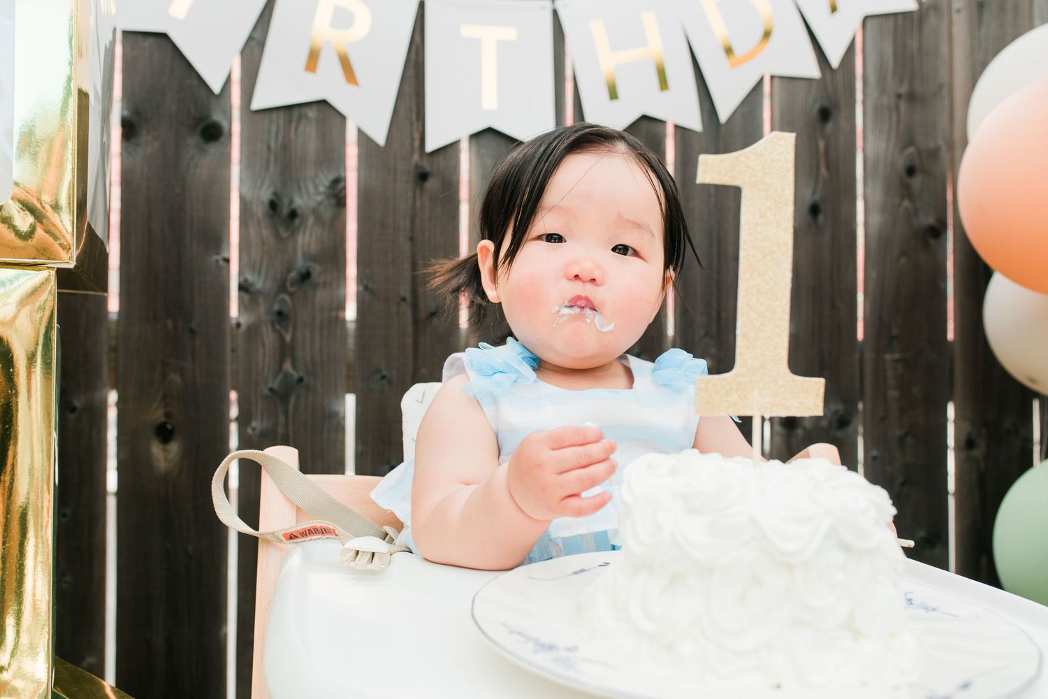 A baby in a white dress sits at a party table decorated with pink and gold balloons and banners.