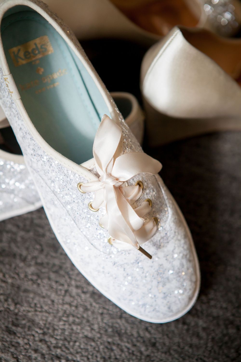 White lace wedding shoes with delicate bow details displayed on a dark surface.