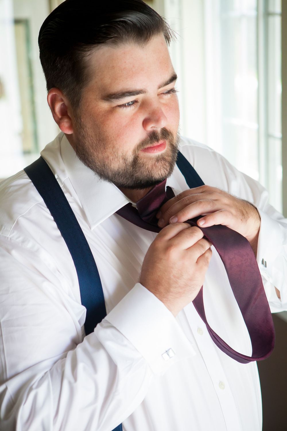 Man adjusts burgundy tie while wearing white dress shirt and navy suspenders.