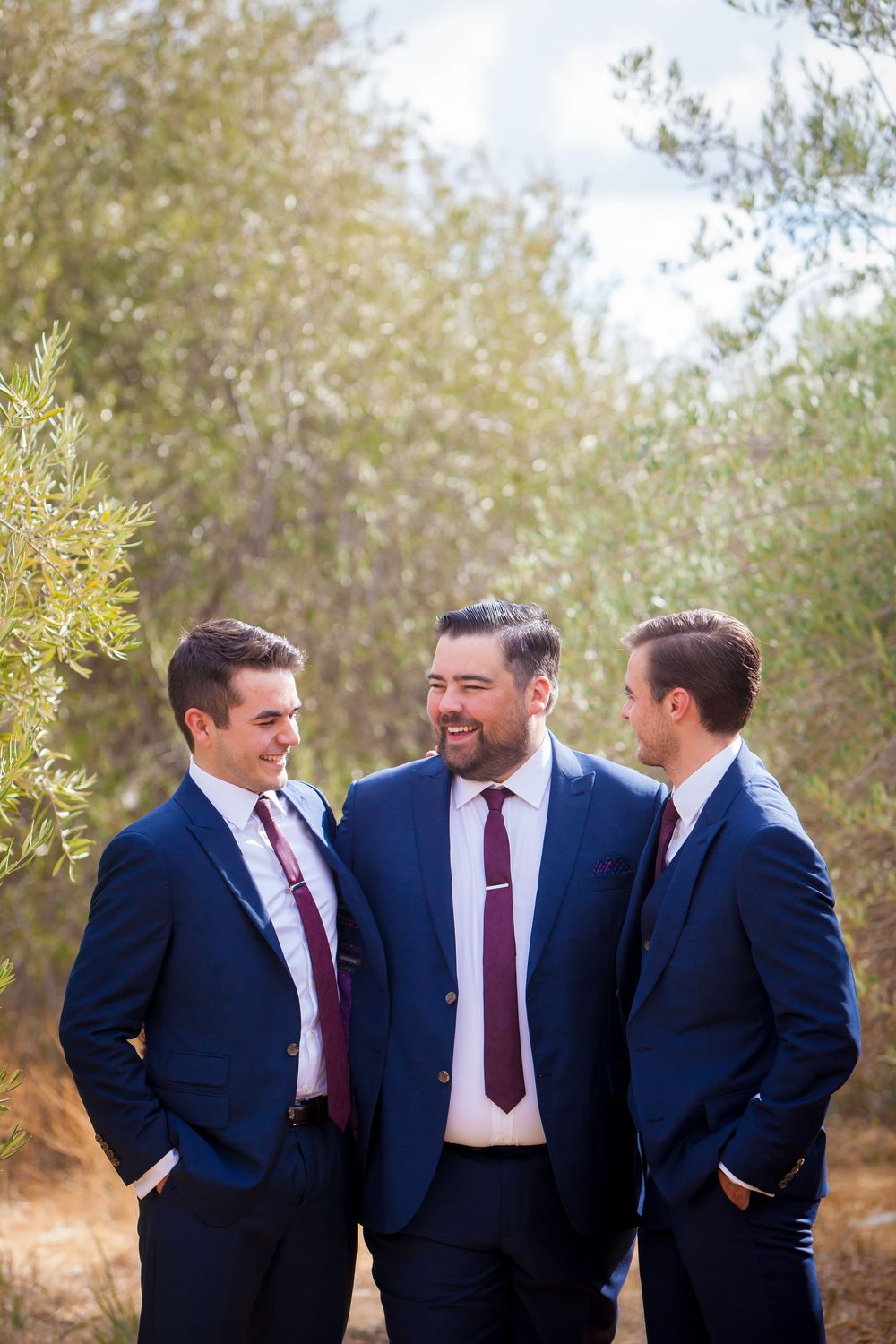 Groomsmen in navy suits and burgundy ties share a laugh outdoors.