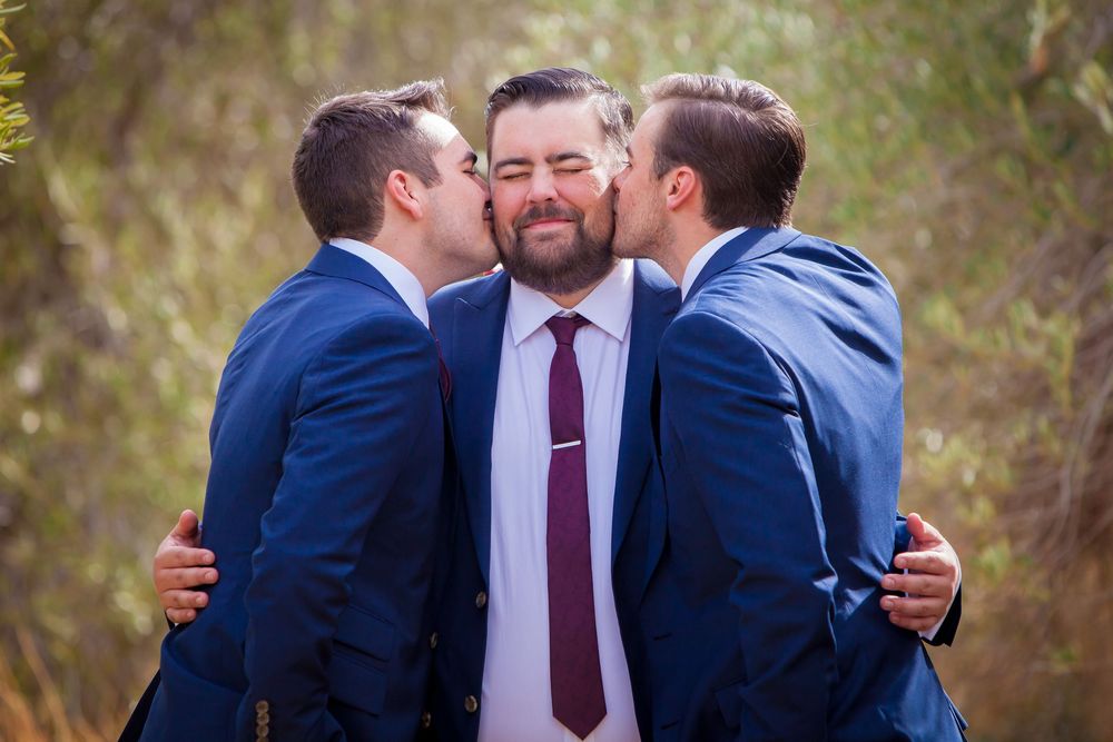 Groomsmen in navy suits and burgundy ties share playful group photos outdoors.