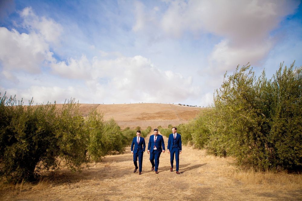 Panoramic desert landscape with blue sky and scattered brush showing distant wedding party figures.