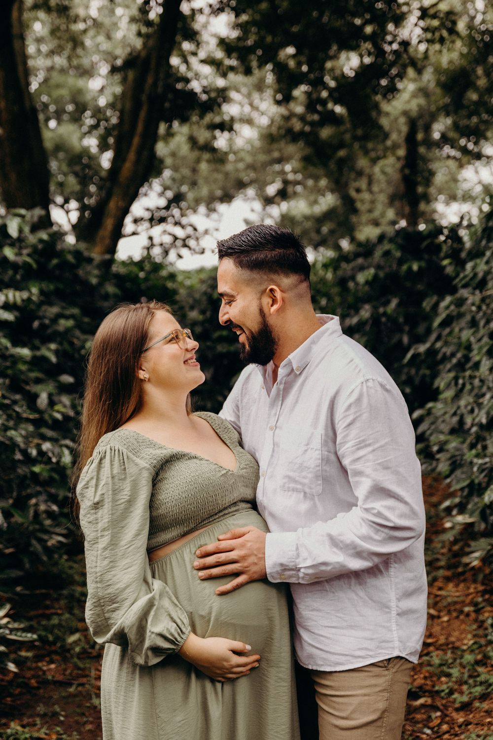 A couple shares an intimate moment in a forest setting as they celebrate their pregnancy in a sage green dress.