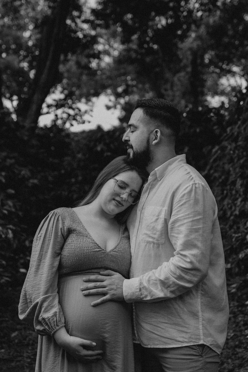 A couple shares an intimate black and white maternity photoshoot moment outdoors among trees.