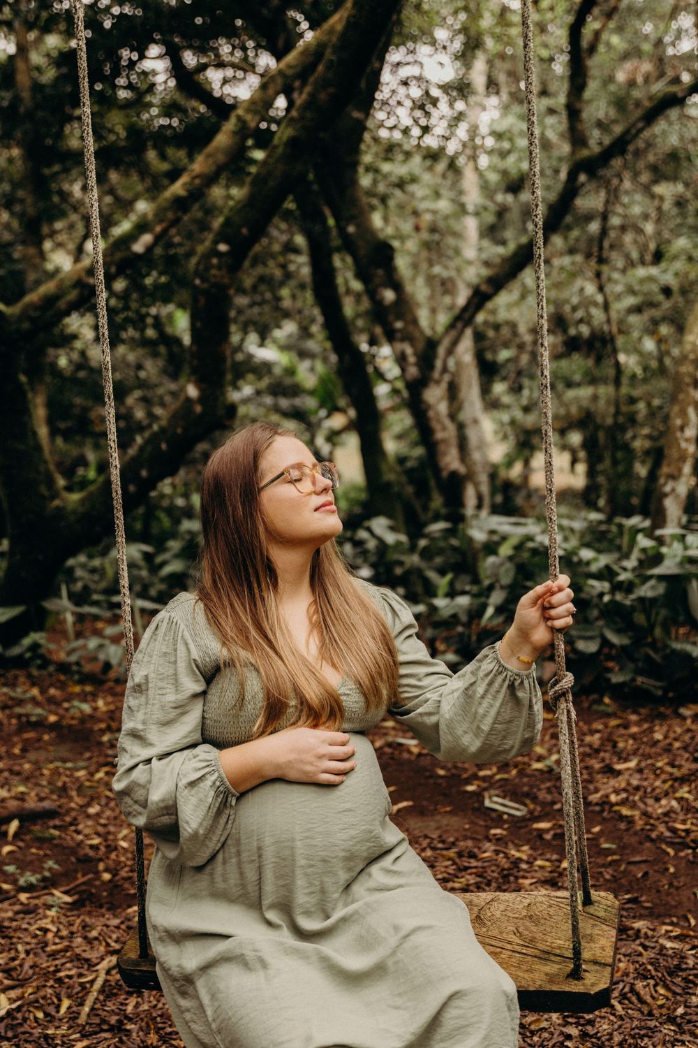 Person in glasses and gray sweater sitting on wooden swing in autumn forest with fallen leaves.