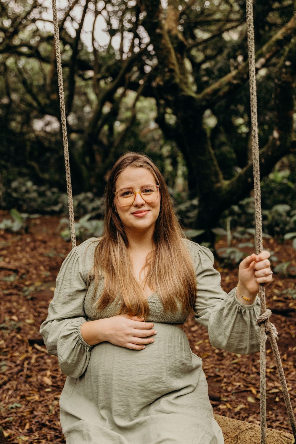 A person in a gray sweater sits on a swing in an autumn forest setting with fallen leaves on the ground.