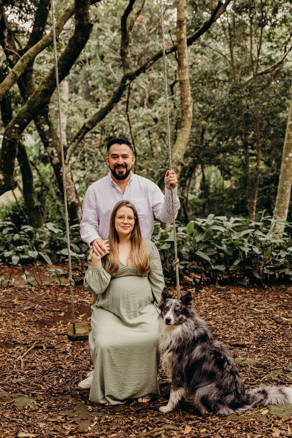 A couple and their dog enjoy a peaceful moment in a wooded setting with fallen leaves.