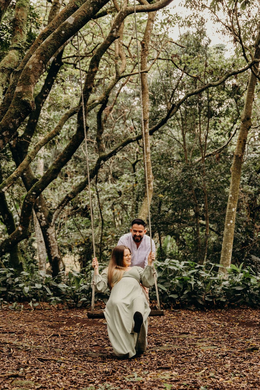 A couple shares a romantic moment while sitting on fallen leaves in a wooded forest setting.
