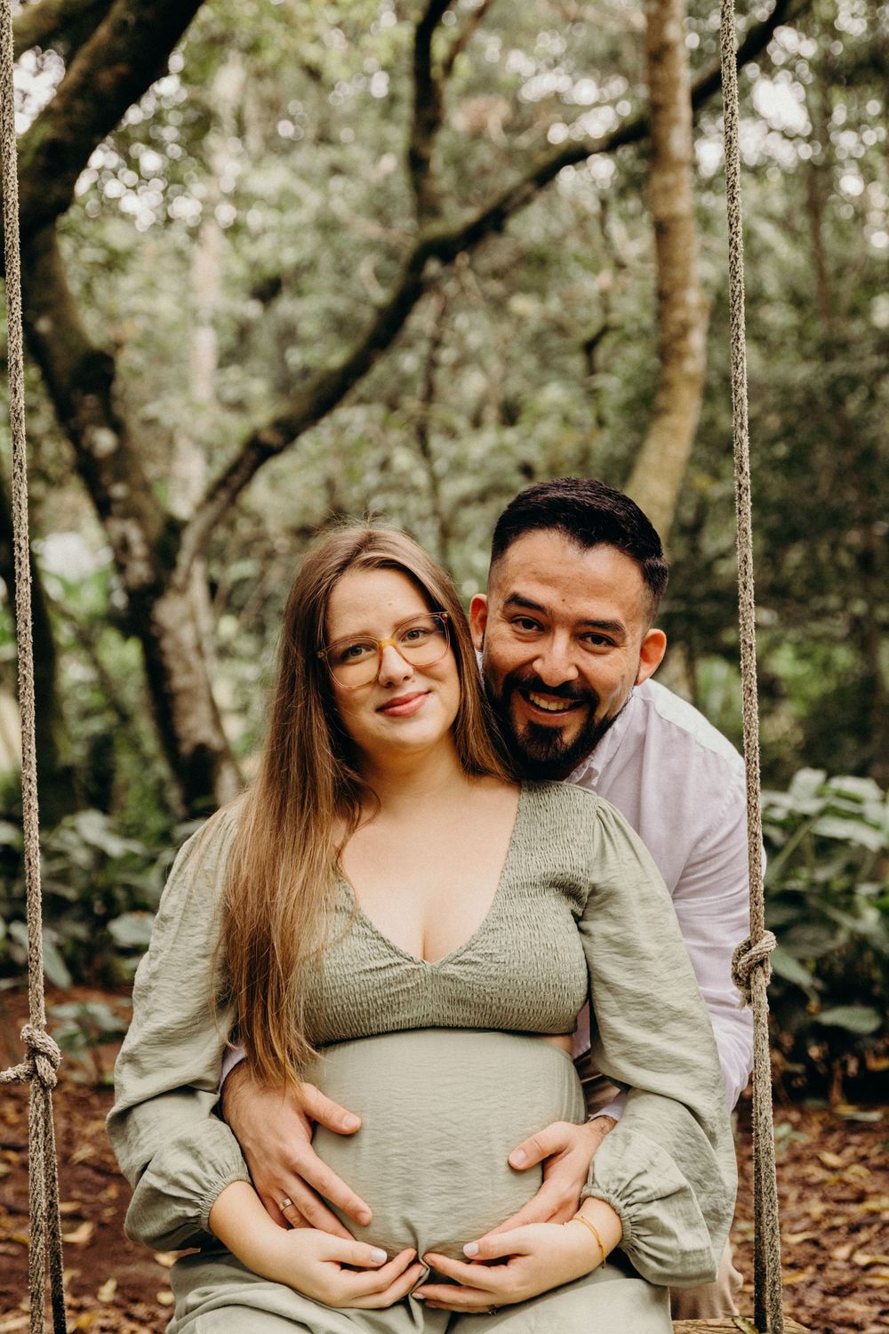A couple embraces while sitting together on a wooden swing in a lush forest setting.