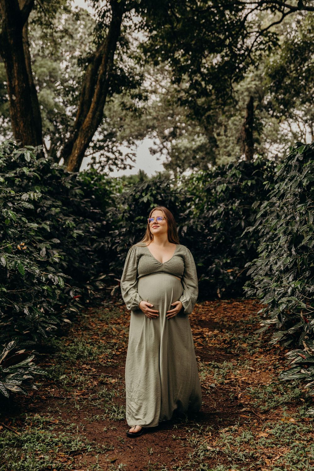A person in an olive green dress poses thoughtfully in a serene forest setting with fallen autumn leaves.