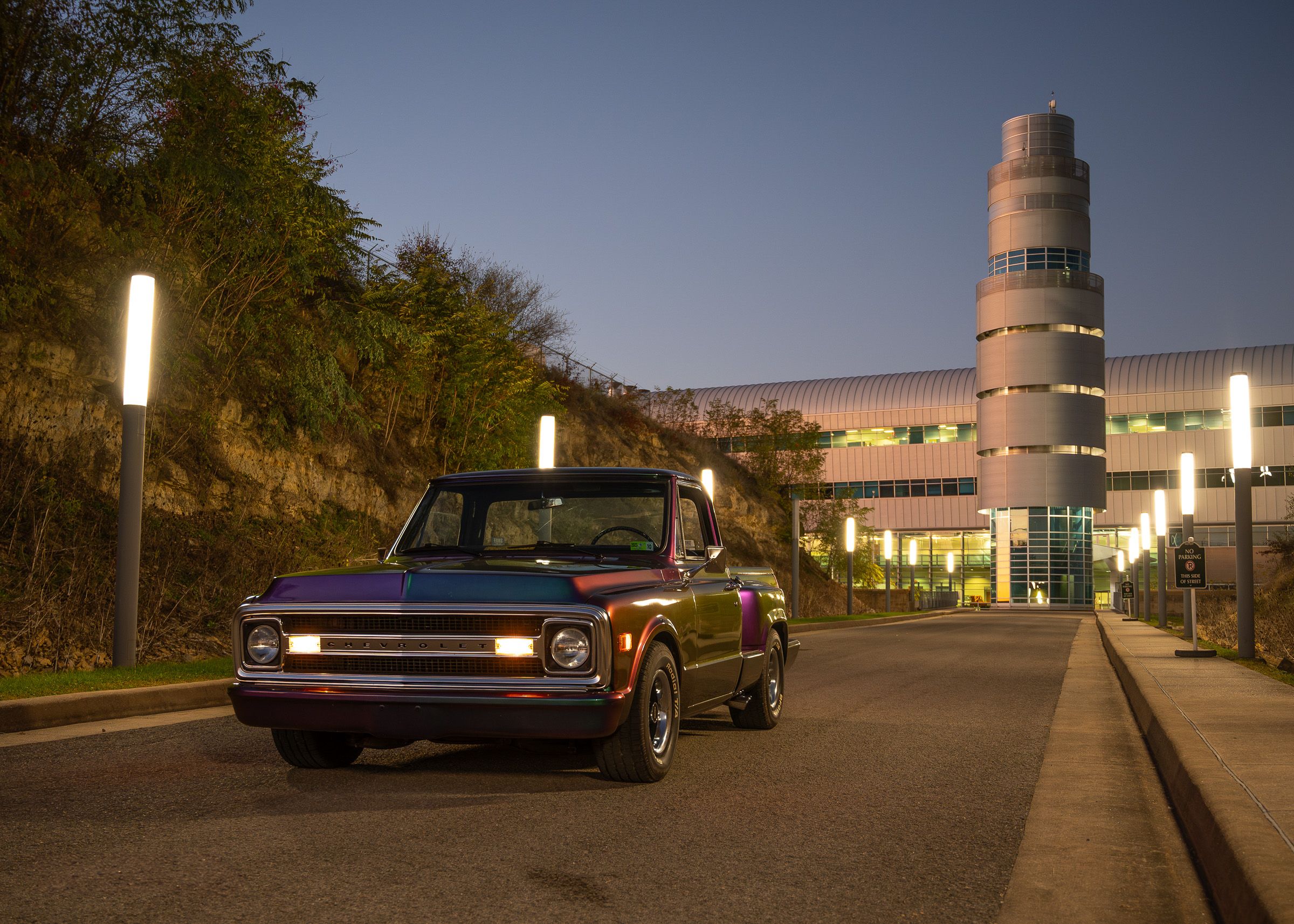 A vintage Chevrolet pickup truck parked at night near a modern building with illuminated architectural features.