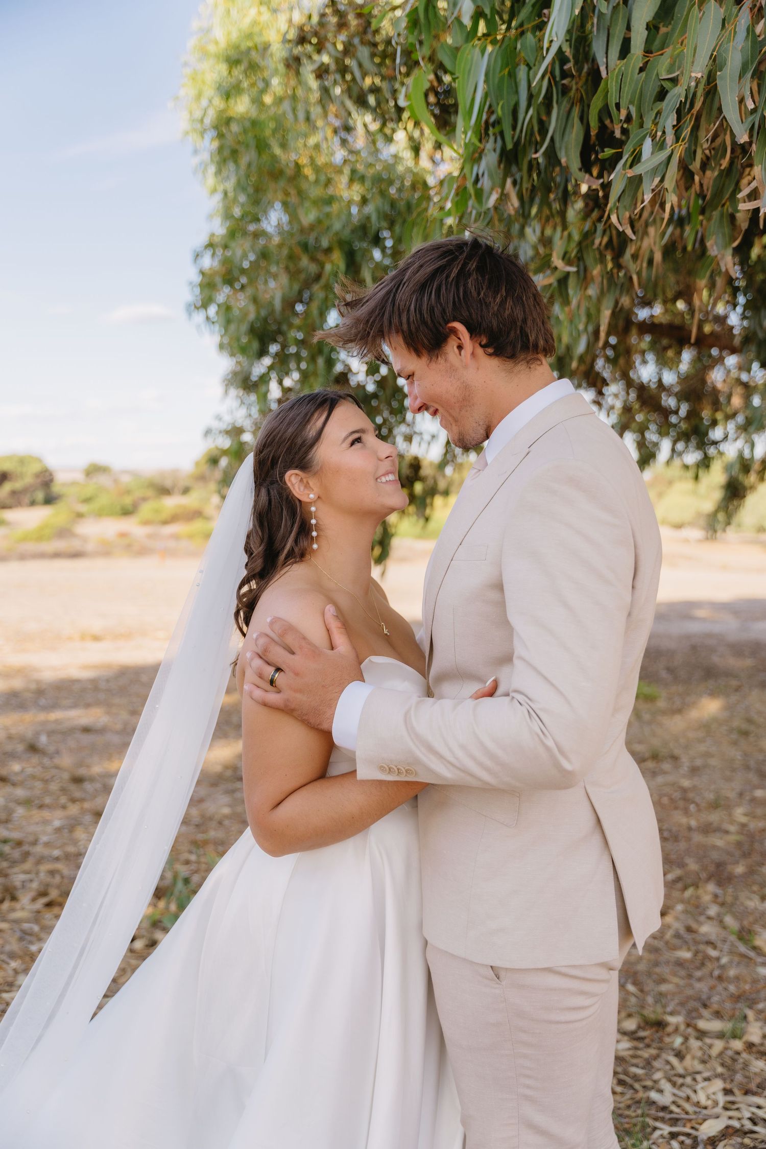 A romantic wedding couple in light beige and white attire share tender moments under eucalyptus trees in a desert setting.