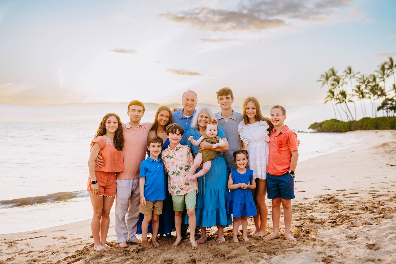 Extended family wearing coordinated blue and coral outfits poses together on a sandy beach at sunset. hawaii family photography kihei wailea