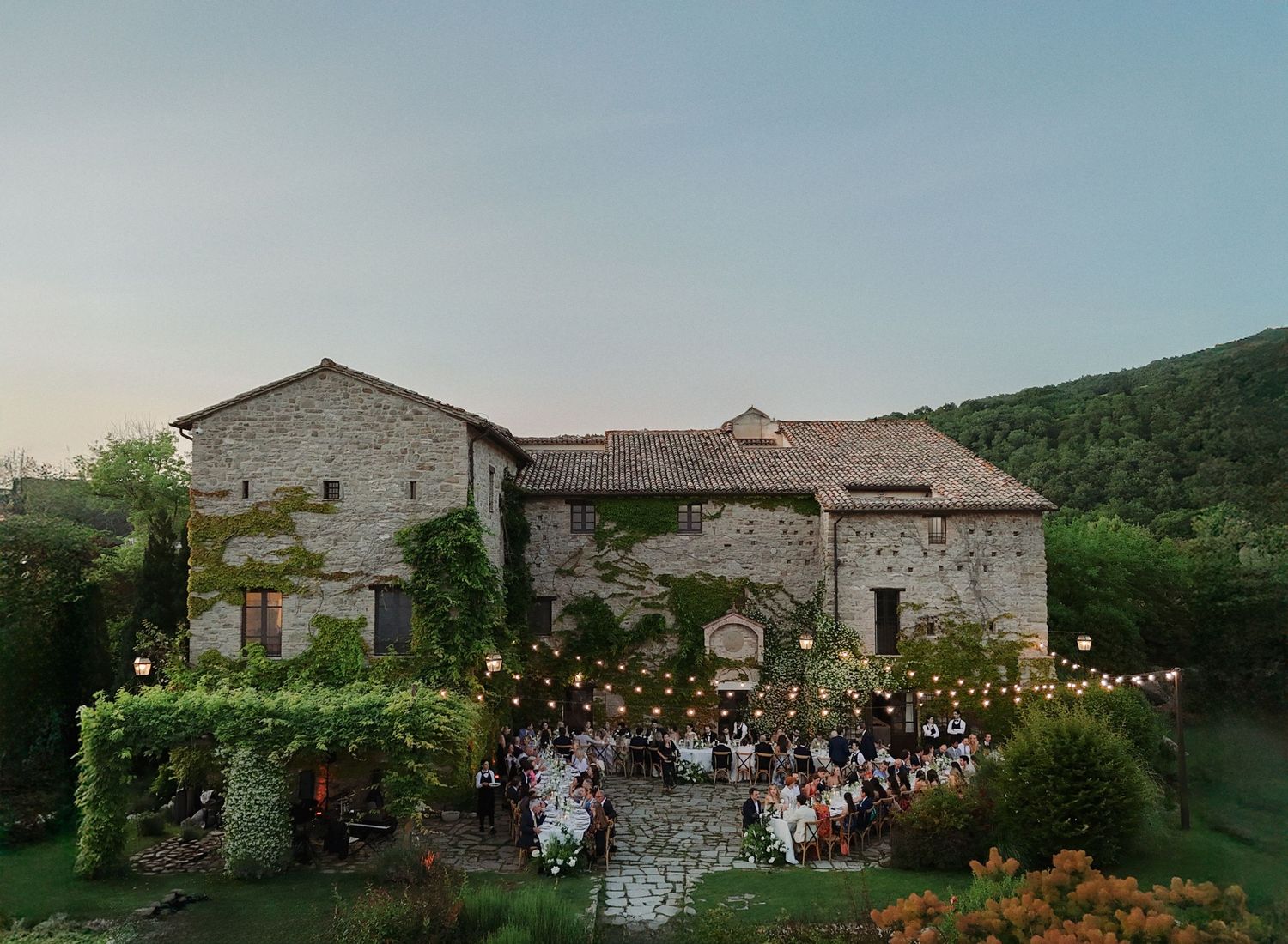 A panoramic view of an outdoor wedding reception at a rustic stone villa with string lights and guests gathered in the garden.