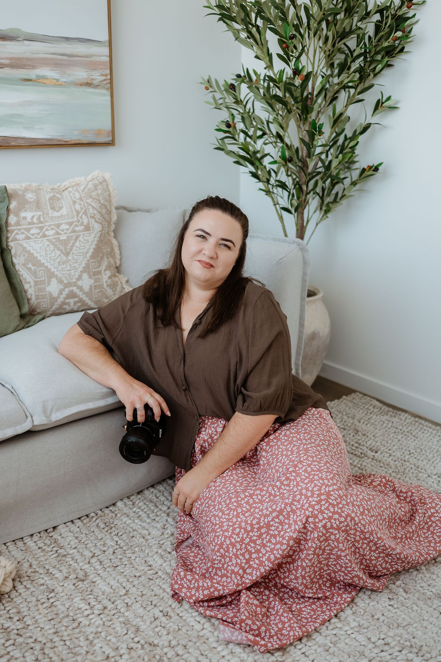 A person in a brown top and floral skirt lounges on a cream rug in a minimalist room with olive plants.