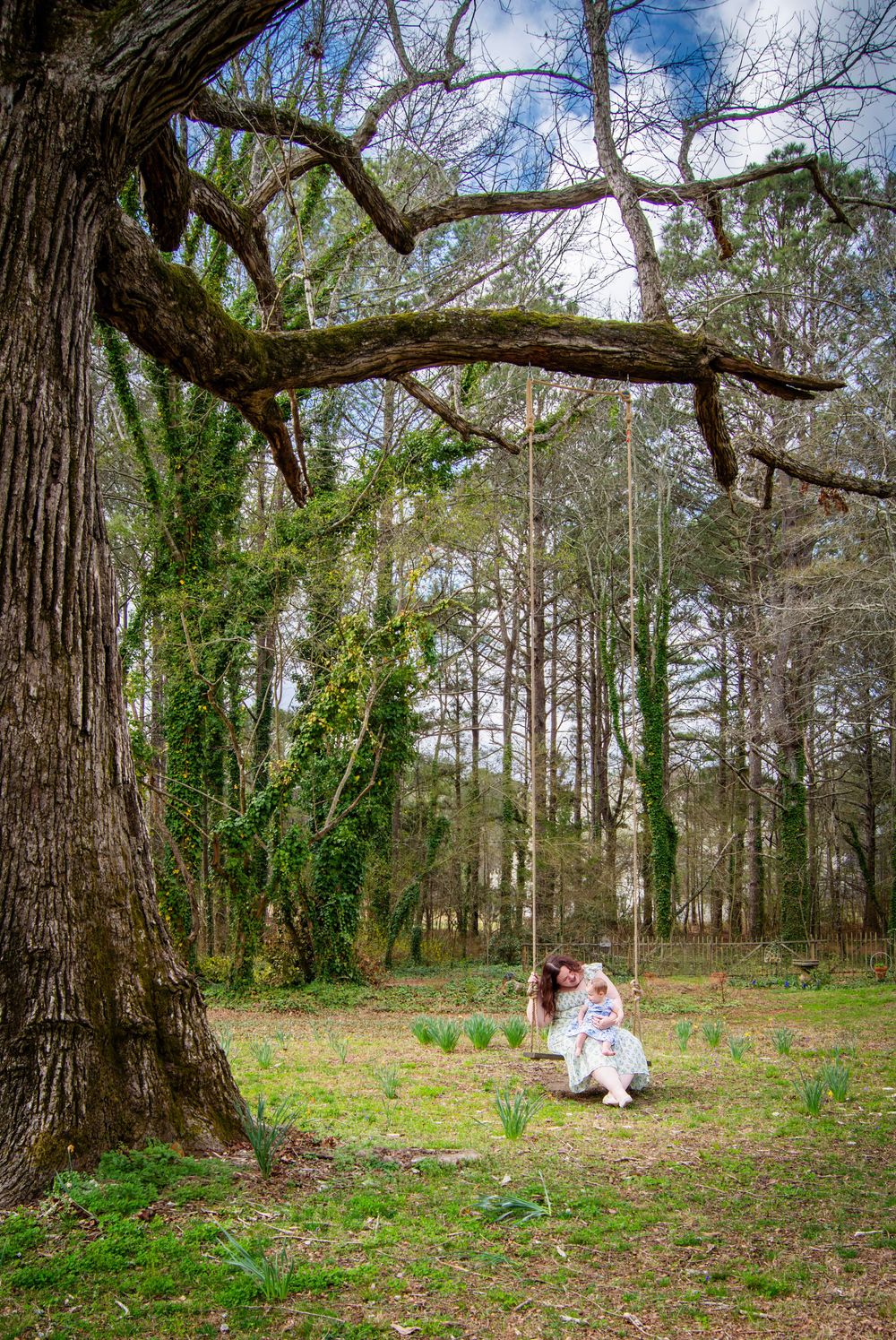 Mother and child sitting together on a rope swing hanging from a large moss-covered tree in a lush green forest.