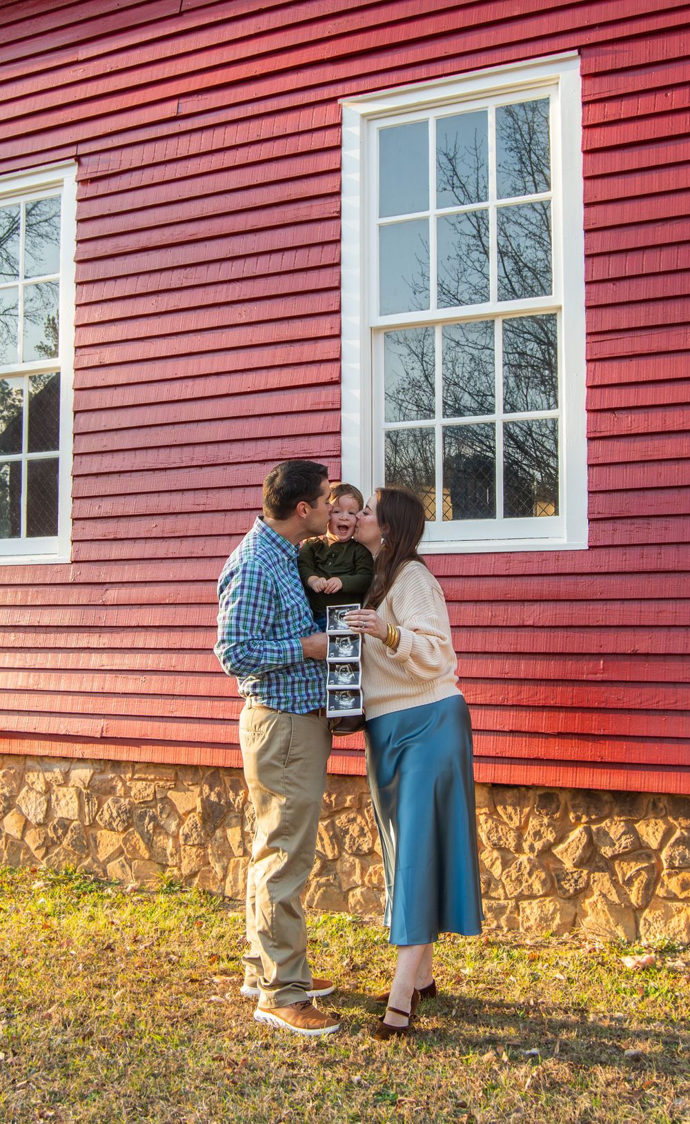A couple kisses while holding their toddler outside a red barn, the woman displaying ultrasound photos for a pregnancy announcement.