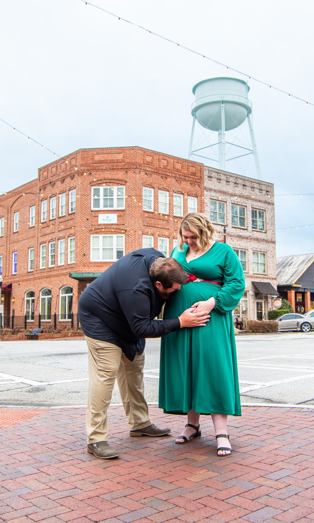Man kissing pregnant woman's belly in green dress outdoors near brick building and water tower.