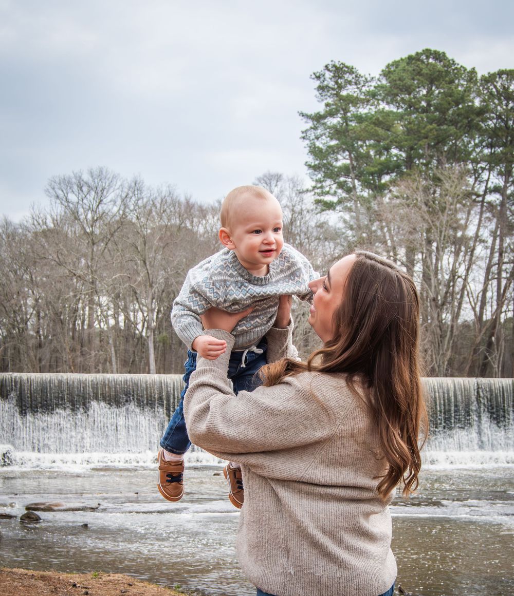 A mother joyfully lifts her baby boy outdoors near a waterfall, both smiling, with bare trees in the background.
