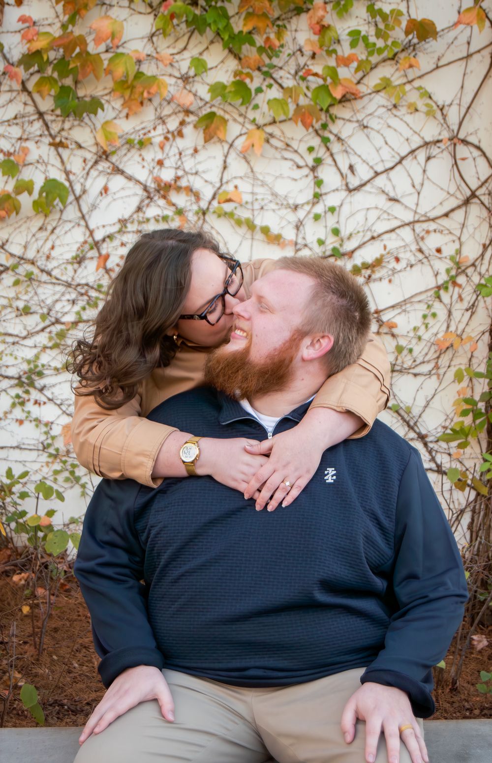 A woman with glasses kisses a red-bearded man outdoors, embracing him warmly against an ivy-covered wall in autumn.