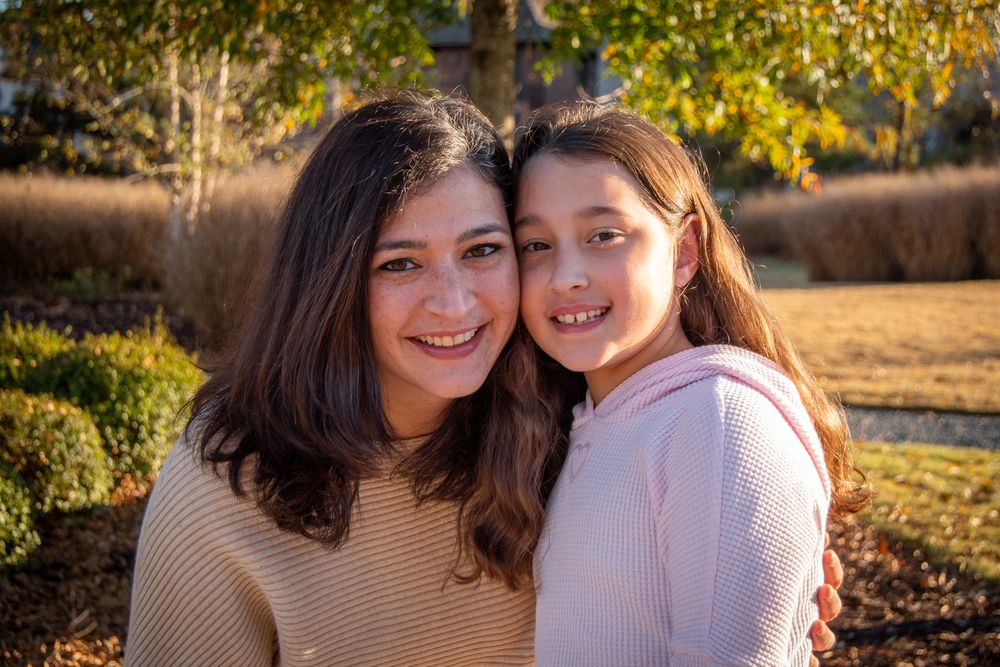 Two people embrace and smile while posing outdoors in autumn with golden sunlight and fall foliage in the background.