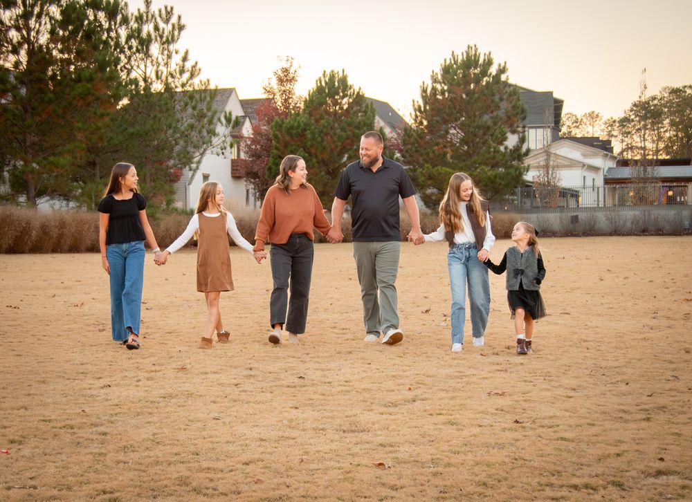 A group walks together holding hands across a sandy field at sunset with pine trees and buildings in the background.