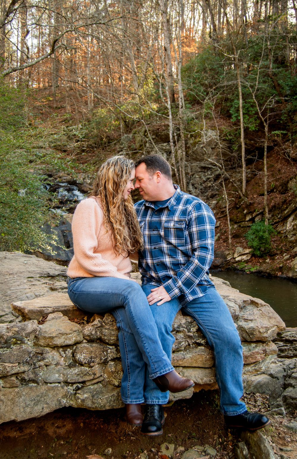 A couple shares an intimate moment while sitting on rocks by a creek in an autumn forest setting.