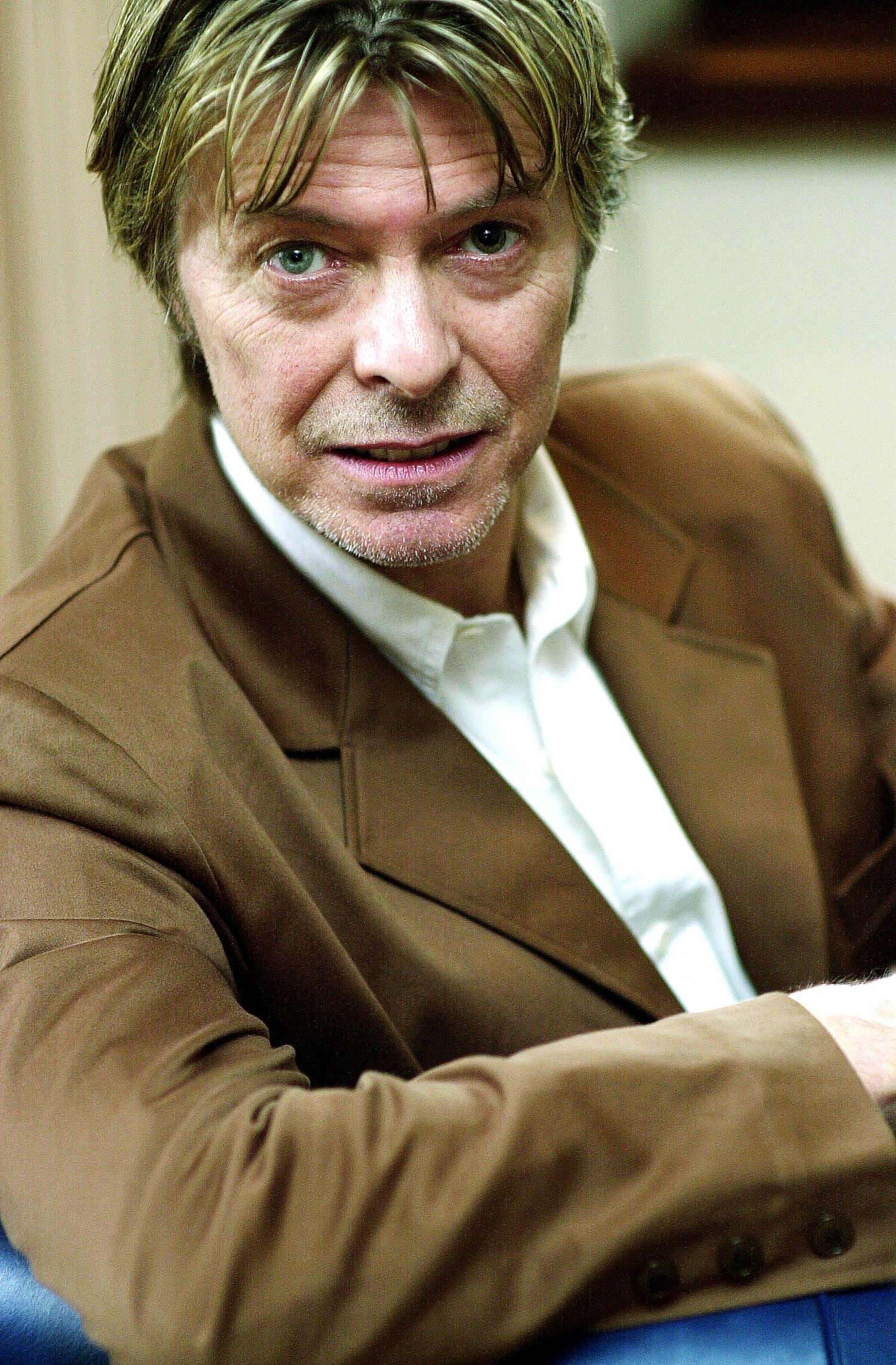 A man in a brown suit jacket and white shirt smiles warmly while seated in a casual pose.