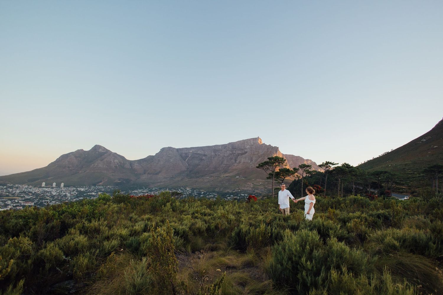 Couple holding hands on a lush hillside with Table Mountain and Cape Town cityscape in the background at sunset.
