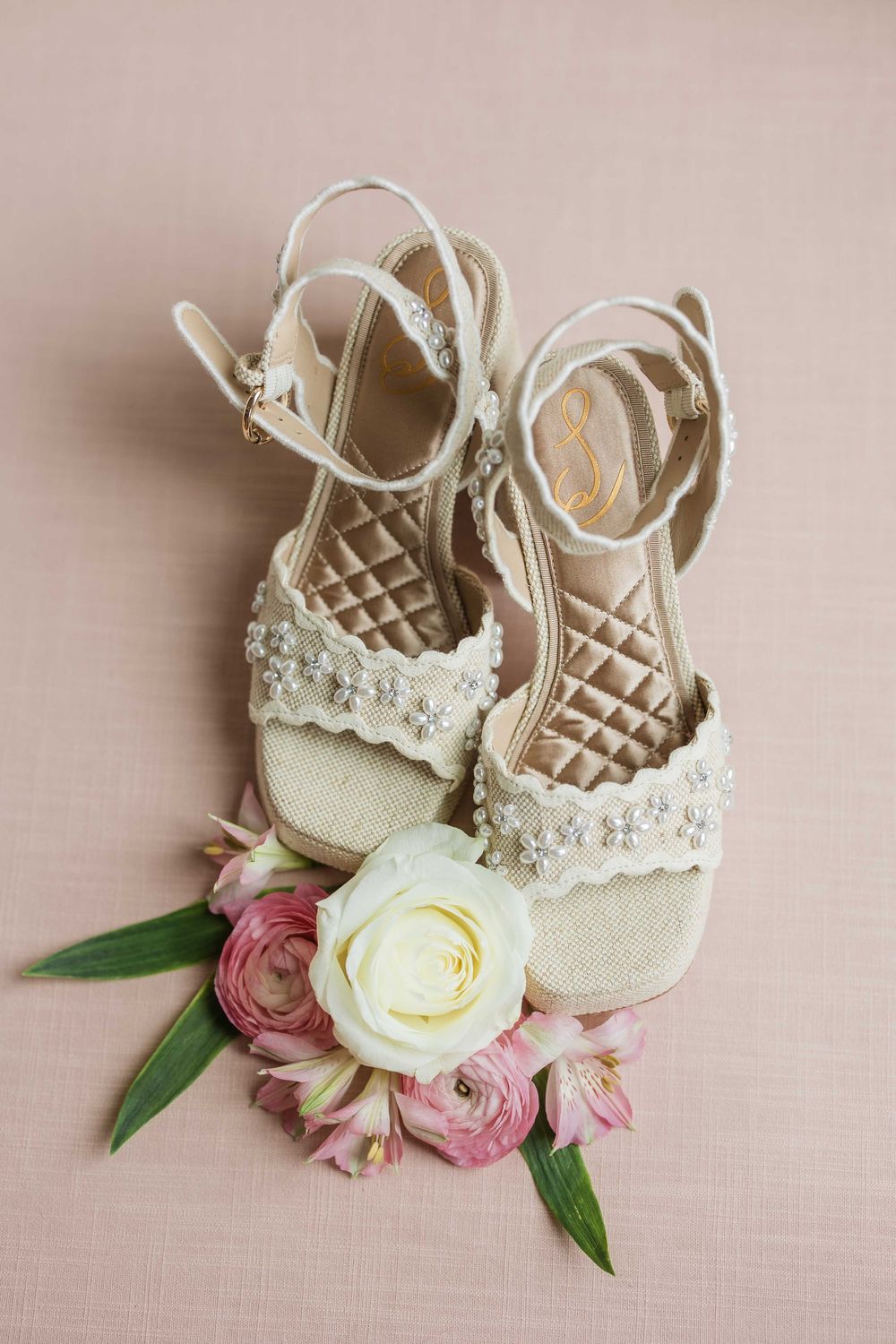 Delicate white beaded baby shoes with pink and cream roses arranged artistically on a blush pink background.
