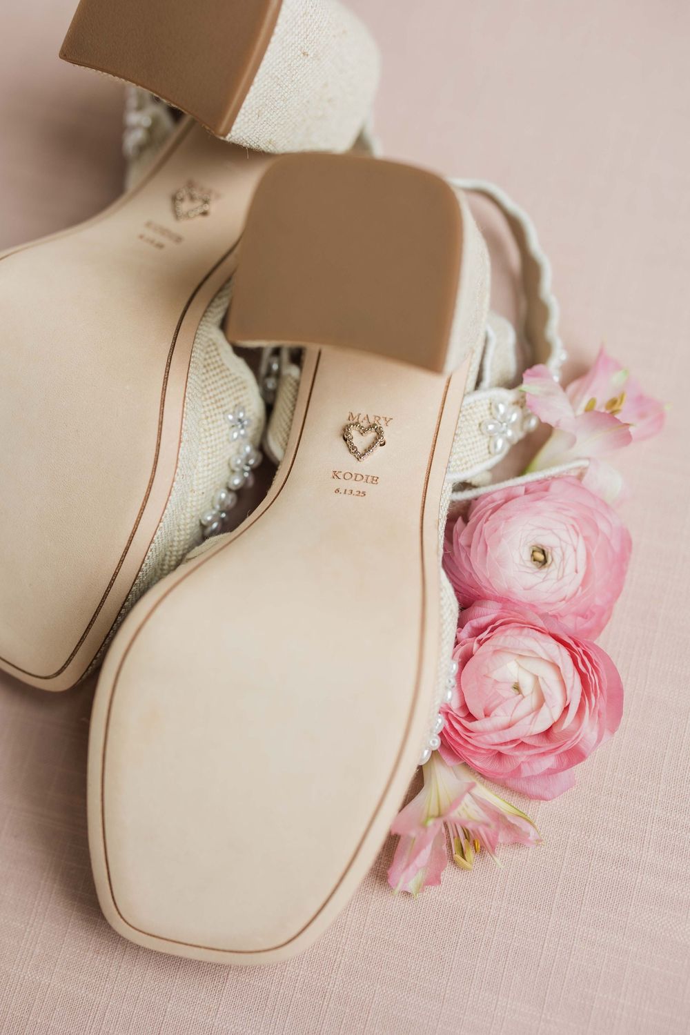 Elegant white bridal shoes with pink ranunculus flowers on a light pink linen background.
