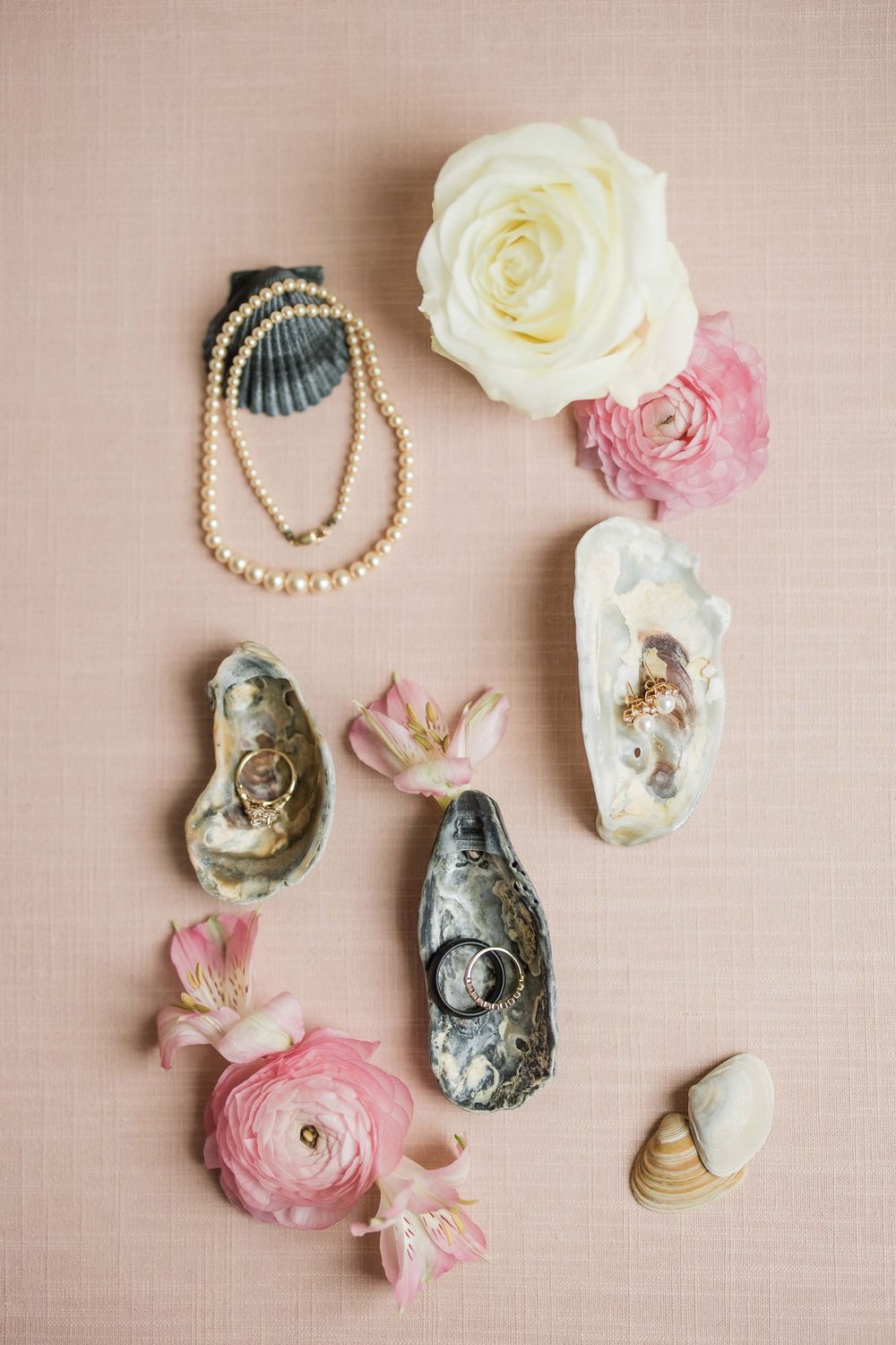 Elegant arrangement of oyster shells, pearl necklaces, and pink and white ranunculus flowers on a blush pink background.