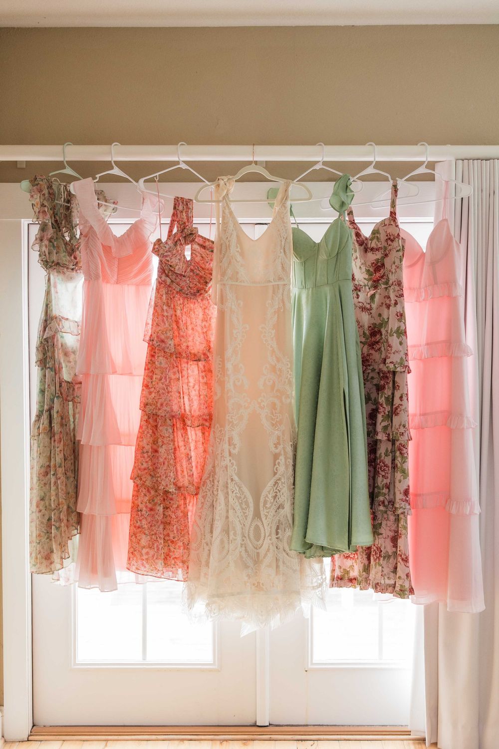 Colorful bridesmaid dresses in coral, peach, sage green and floral prints hanging by a window in natural light.