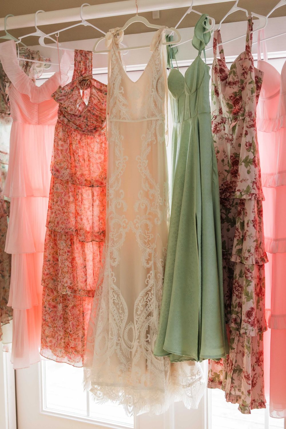 A collection of vintage-style dresses in pink, green, and floral patterns hanging on a clothing rack against a white wall.