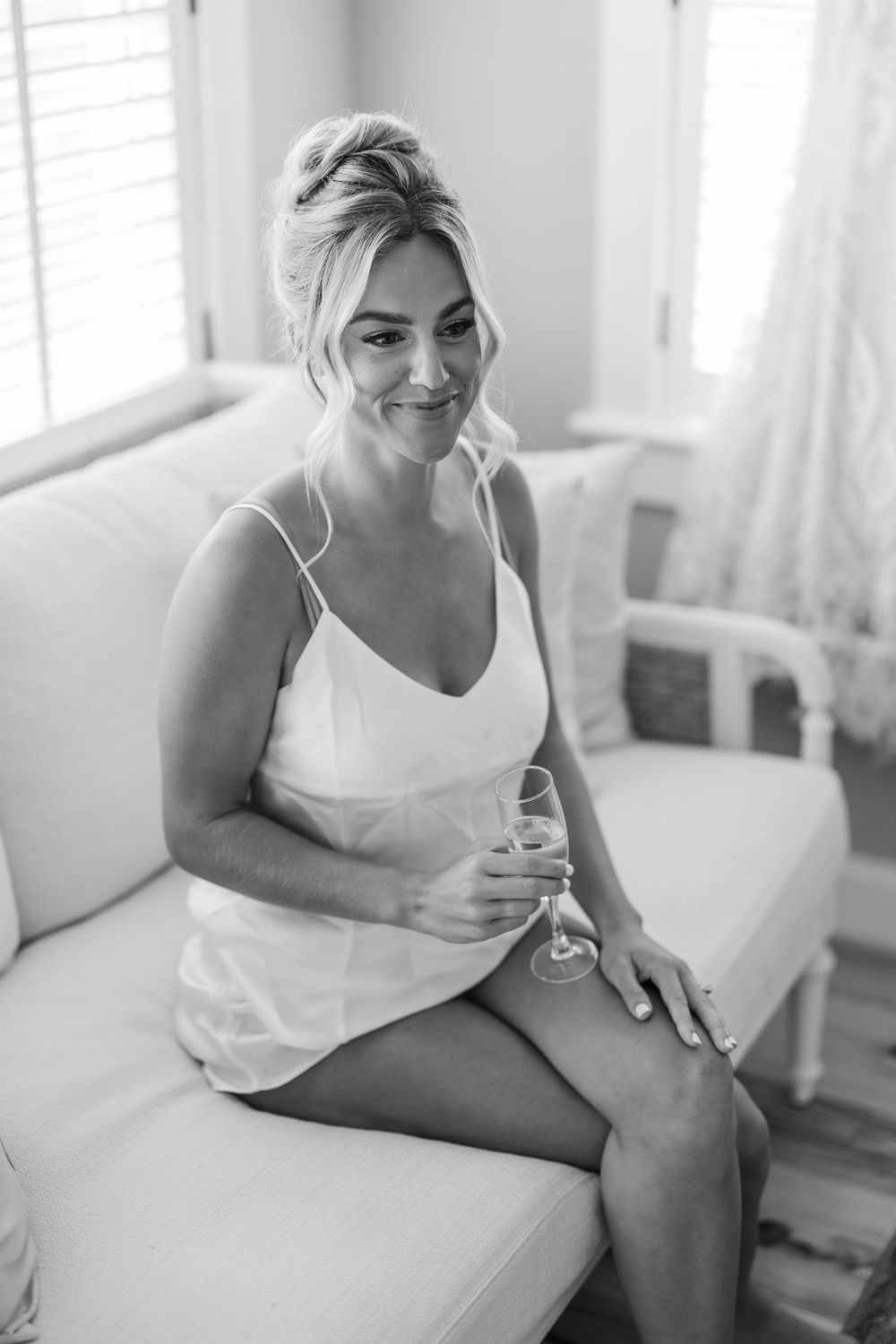 A person in a white tank top sits casually on a couch in a bright sunlit room with window blinds.