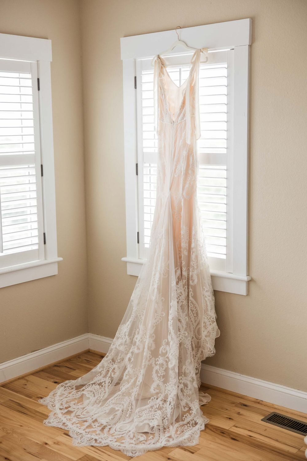 A delicate lace wedding dress with a long train hangs elegantly by a window with white shutters in a sunlit room.