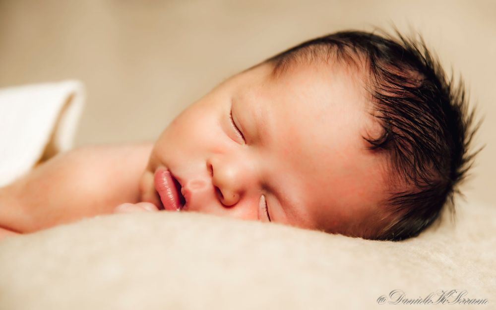Peaceful sleeping newborn baby with dark hair resting on a soft white blanket.
