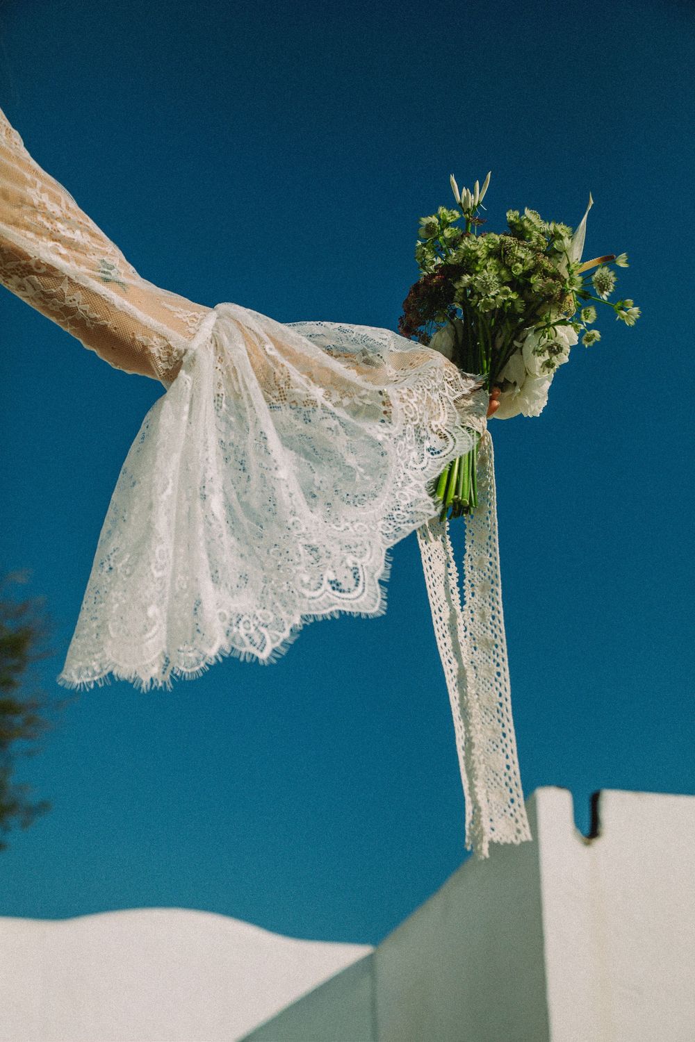 White lace wedding dress sleeve details flutter against a bright blue sky in an artistic photo series.