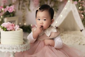 Close up of a baby girl eating a handful of birthday cake. Background has a white tent with hanging greenery and pink flowers.