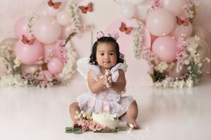 Baby girl sitting and smashing a birthday cake with a light pink wildflower backdrop with balloons and orange butterflies
