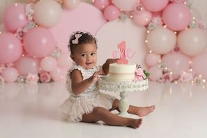 baby girl sitting with her birthday cake in front of a wall of light pink balloons and flowers, she is wearing a white lace dress