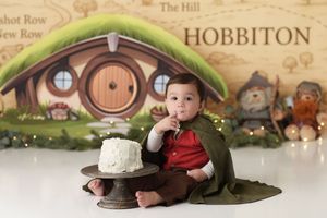 Baby boy with a Hobbit backdrop and costume with a cape, feeding himself a bite of cake.