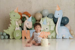 One year old birthday boy sitting with his cake in front of a pastel dinosaur themed setup with balloons in blues, greens and orange.