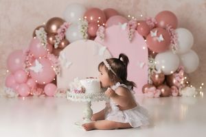 baby girl leaning in to take a bite out of her birthday cake face first with a balloon backdrop in shades of pinks